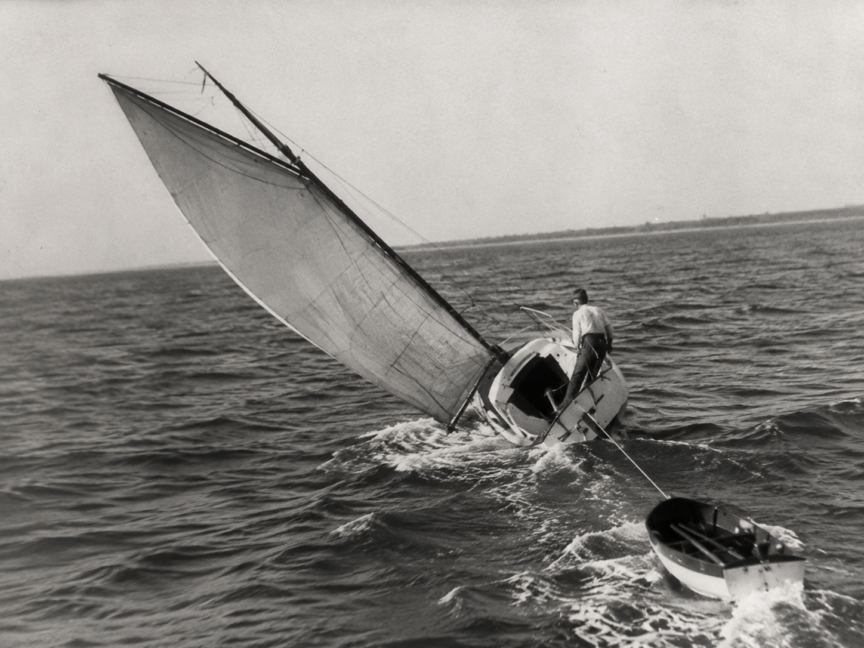 Vintage black-and-white image of a sailboat heeling in strong wind while towing a dinghy