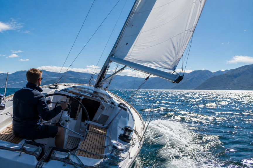 Photorealistic view from the cockpit of a modern fiberglass day sailer sailing on a beam reach under mainsail alone, with the skipper steering from behind the wheel and looking ahead across sparkling blue water toward a mountainous coastline.