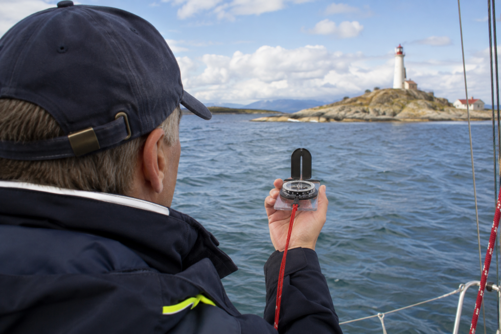 Sailor using a hand-bearing compass to take a bearing on a lighthouse while navigating a sailboat along the coast.