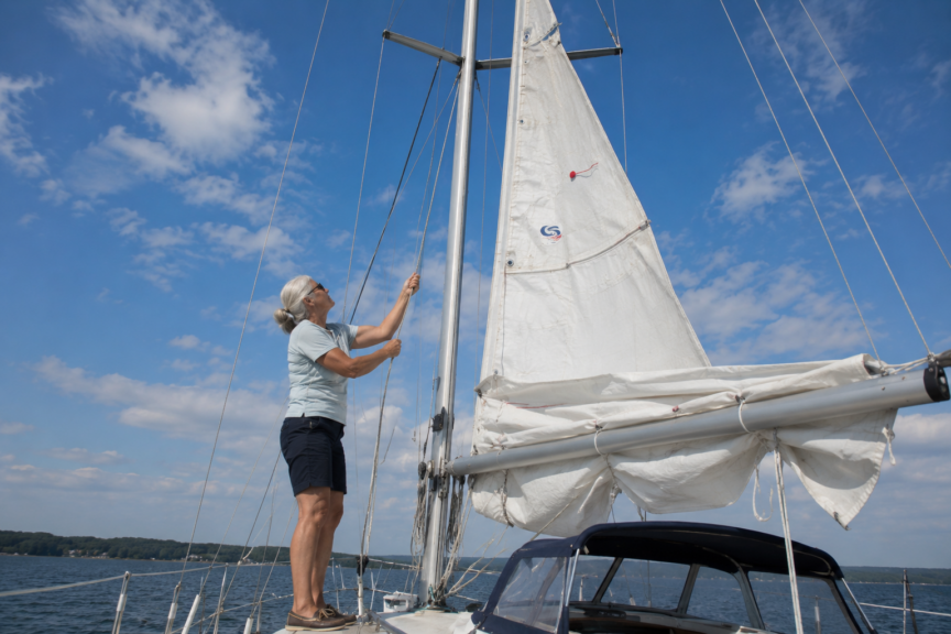 A beginner sailor stands at the mast pulling the main halyard as the mainsail is being hoisted. The sail is about three-quarters raised, with loose folds still resting on the boom during the hoisting process.