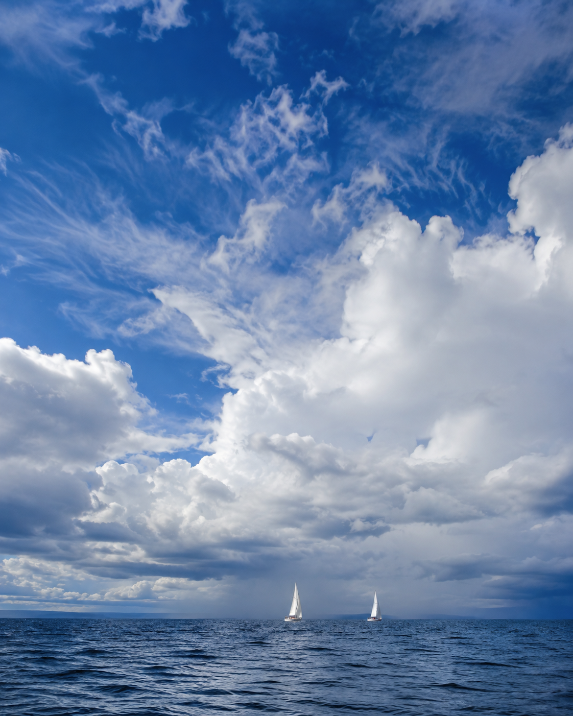 Two small sailboats beneath towering clouds on a vast expanse of open water